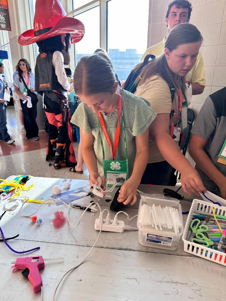 A 4-H participant works on a hands-on craft and engineering activity at a workshop table during a statewide 4-H event. The table is covered with craft supplies, including pipe cleaners, pom-poms, scissors, markers, glue sticks, and a hot glue gun. The participant is assembling materials while wearing a 4-H name badge and event lanyard. Other attendees stand nearby working on projects, and Oklahoma State University's mascot, Pistol Pete, is visible in the background. Large windows provide natural light to the activity area. The photo highlights creativity, problem-solving, and experiential learning through 4-H educational programs.