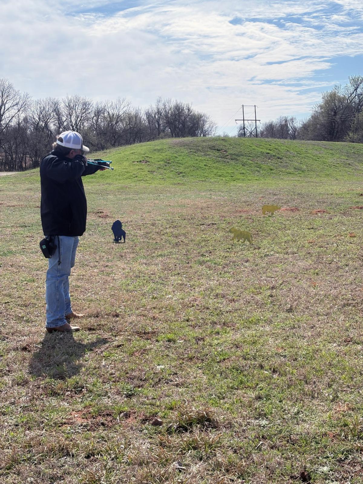 Person wearing a white cap, dark jacket, jeans, and boots stands in an open grassy field and aims a slingshot toward three animal-shaped metal silhouette targets positioned at varying distances. A low green hill rises behind the targets, with trees, utility poles, and power lines along the horizon beneath a bright, partly cloudy sky.