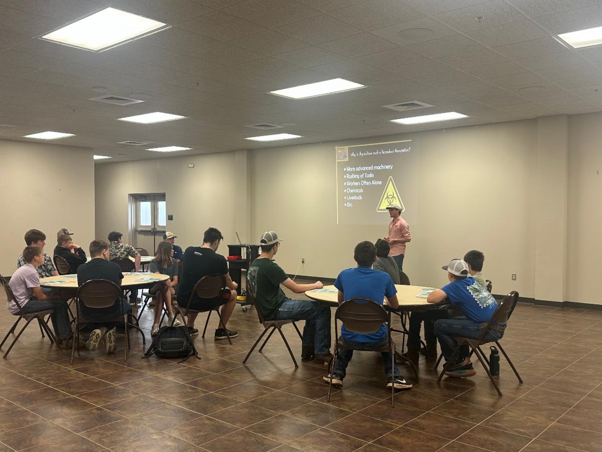 Youth seated at round tables in a classroom, listening to a presenter standing at the front next to a projected slide about agricultural safety that lists hazards such as machinery, chemicals, and livestock.
