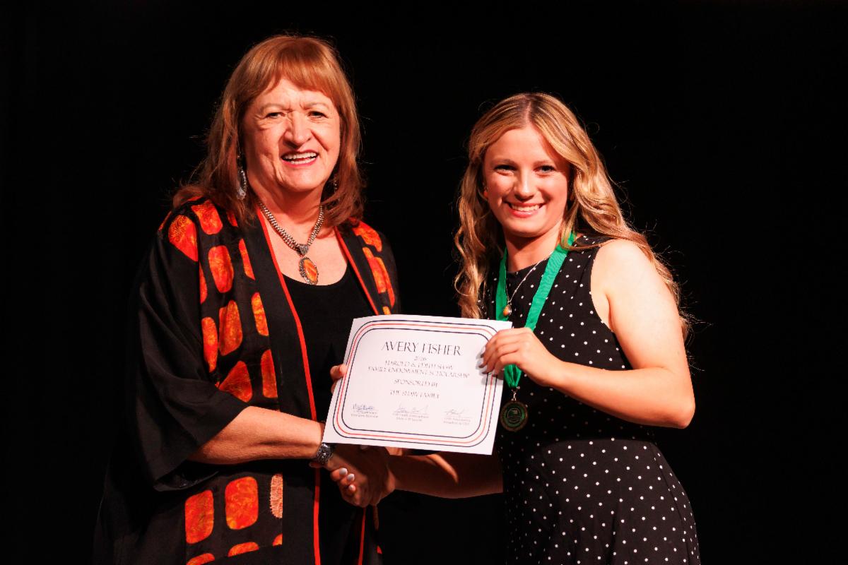 Avery Fisher receives a scholarship certificate during the 105th Oklahoma 4-H State Roundup. Fisher and a presenter pose on stage while holding the certificate and shaking hands.