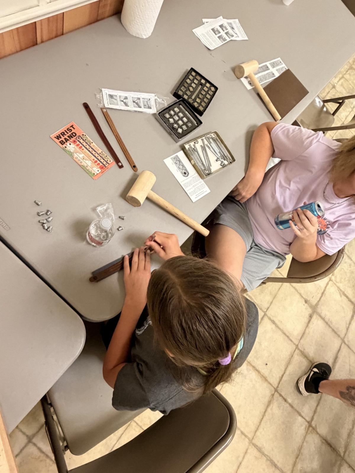 Youth participants work on leathercraft projects at a workshop table. Leather wristbands, stamping tools, wooden mallets, and instruction sheets are arranged on the table as one participant stamps a design into a leather bracelet.