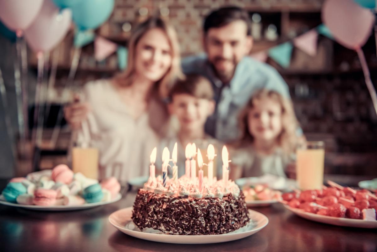 A birthday celebration scene featuring a chocolate cake topped with several lit candles on a table in the foreground. Plates of colorful desserts, including macarons and red sweets on skewers, sit beside glasses of a light-colored beverage. Pastel balloons and decorative bunting are visible in the background. Four people are seated behind the cake, intentionally blurred, while the cake and candles remain in sharp focus. Warm indoor lighting creates a festive atmosphere.
