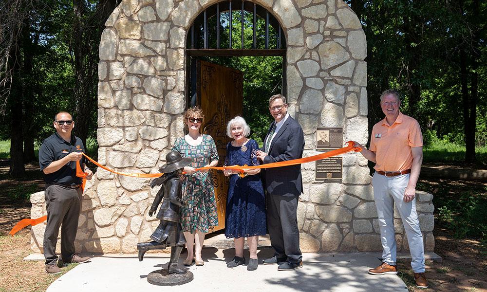 Five people stand in front of a stone arch and hold an orange ribbon to be cut for a grand opening.