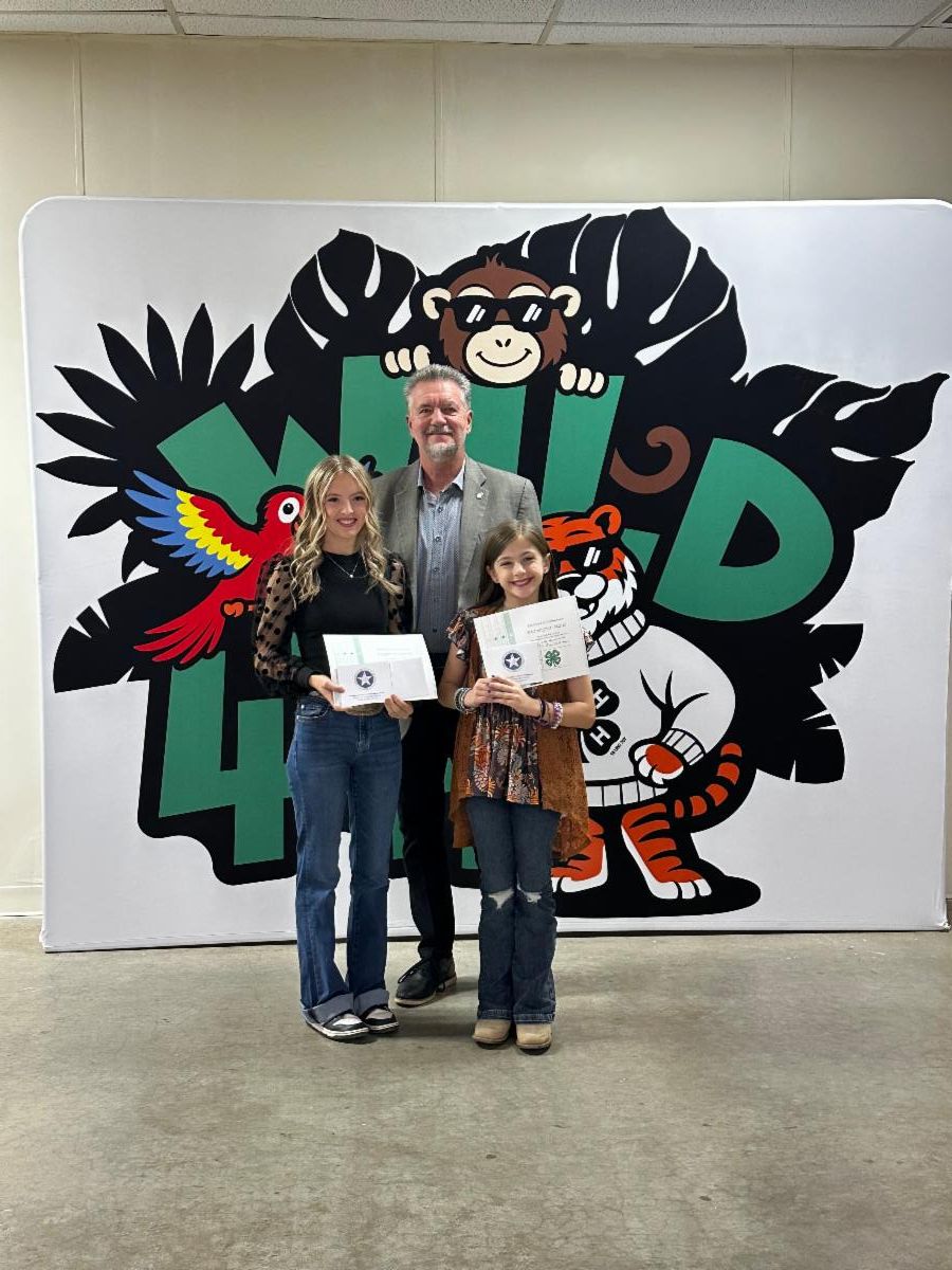 Three individuals stand in front of a large colorful 4-H-themed backdrop featuring cartoon animals and oversized green “4-H” lettering. Two youth stand in the front holding framed certificates, while an adult stands behind them. The group is posing indoors on a concrete floor. The backdrop includes a red parrot, a monkey wearing sunglasses, and a tiger wearing a 4-H scarf. All three individuals are facing the camera and holding or displaying awards received during a 4-H recognition event.