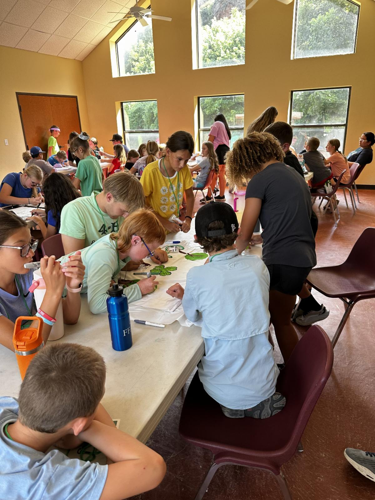 Youth participants work together on a 4-H craft project, decorating T-shirts with 4-H clover designs during a group activity in a bright classroom setting.