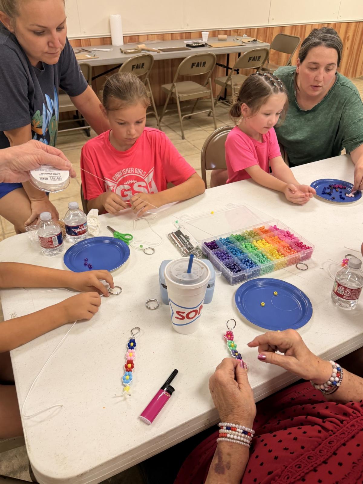 Participants of different ages sit around a table making colorful beaded keychains. Containers of rainbow-colored beads, string, scissors, and finished bead creations are spread across the table as adults assist children with the craft activity.