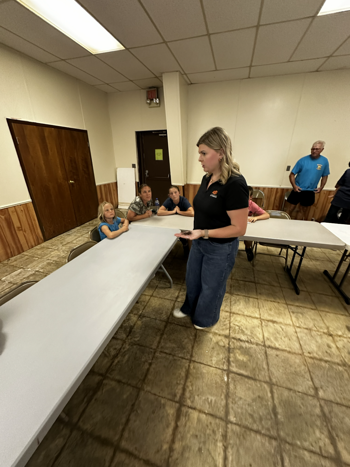 An educator presents an insect specimen during a 4-H entomology workshop while youth and adults participate in an interactive science learning experience.
