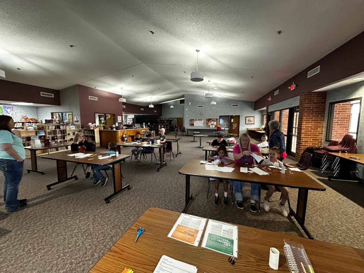 Children and adults are seated at several tables throughout a large, open community room, working on paper craft projects using scissors, paper strips, and small supplies. An adult at the front table helps two children assemble their materials, while other