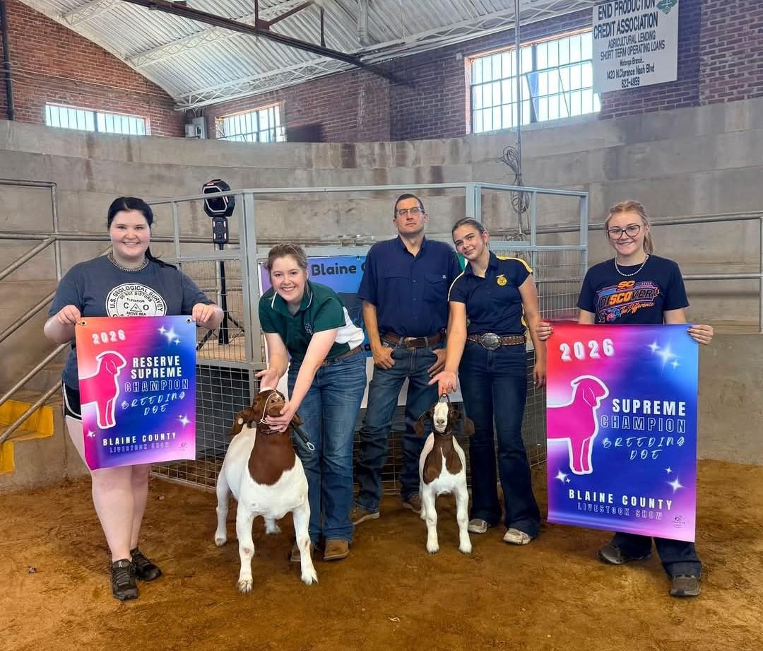 Livestock show winners pose in a show arena at the Blaine County Fair alongside two Boer goats. Four youth exhibitors and one adult stand behind the animals, while two exhibitors hold large award signs reading "2026 Reserve Supreme Champion Breeding Doe" and "2026 Supreme Champion Breeding Doe." The photo was taken inside the livestock barn, with show pens and arena fencing visible in the background.