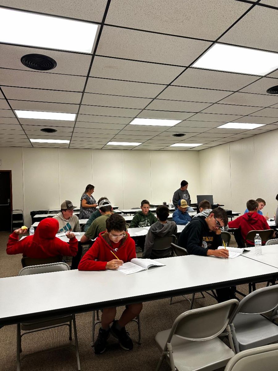 Youth seated at tables in a classroom setting, working on papers and writing in notebooks while two adults stand nearby, providing instruction or assistance.