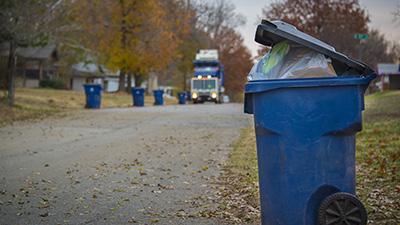 A recycling bin on the side of the road with a recycling truck moving toward it.