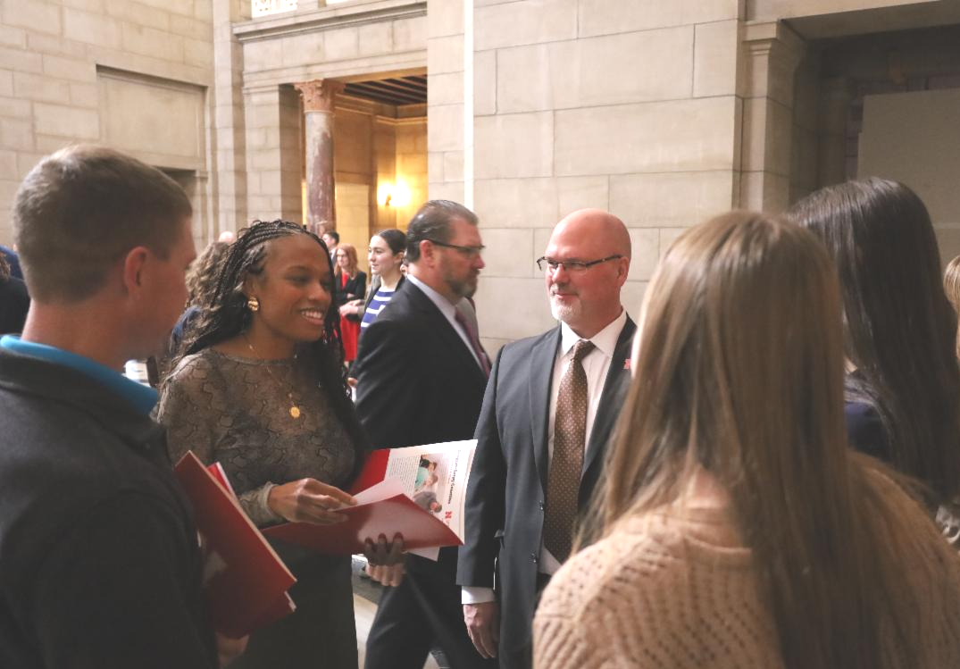 A group of adults in business attire stand and talk inside a large stone building with columns and high ceilings. Several people are holding red folders or papers while conversing in small groups.