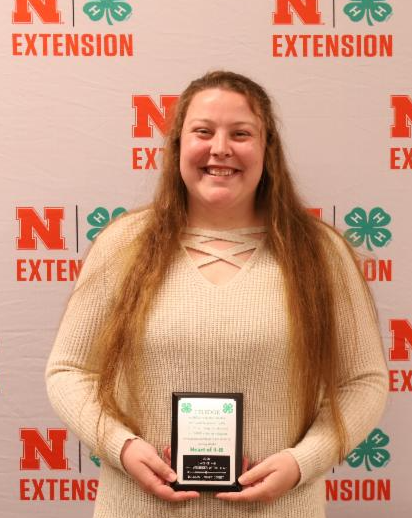 woman holding a plaque, standing in front of a sign that says NE Extension and has 4-H clovers