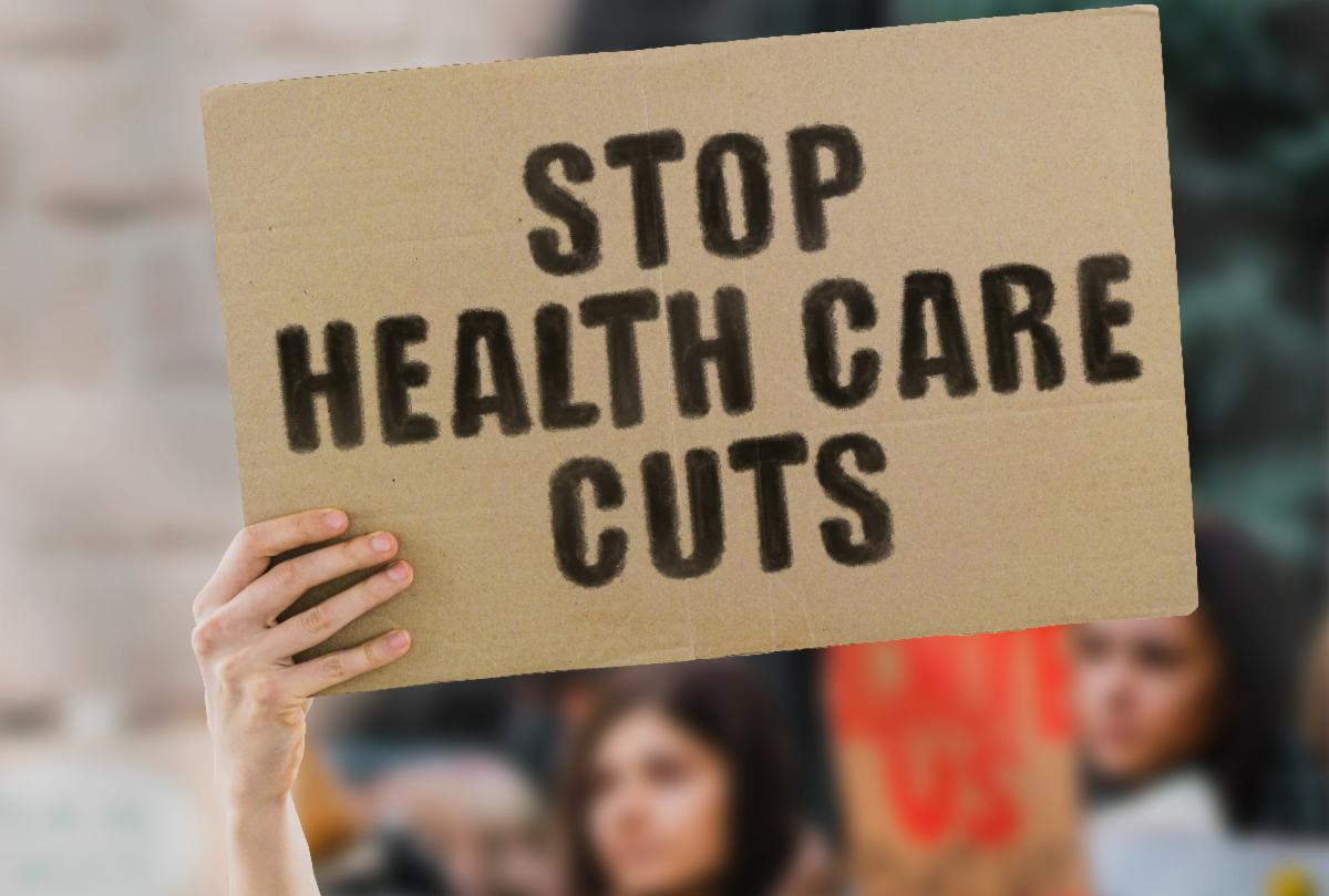 a brown sign with large black letters STOP HEALTHCARE CUTS held up by a hand, blurred people in a crowd in the background