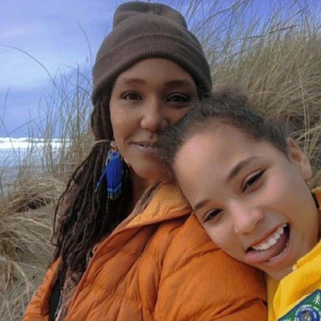 a smiling woman with a beanie covering long dark braids, she is wearing a puffy orange jacket. Her daughter is lying her head on her shoulder smiling with her tongue out. She is wearing a yellow jacket. There are tall brown grasses in the background with water and a blue grey horizon