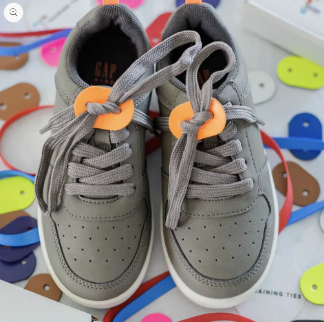 Training Ties Shoe Tying Tool for Kids- a pair of grey kid's shoes with an orange , leather disk at the base of the laces, just above the tongue of the shoes. 