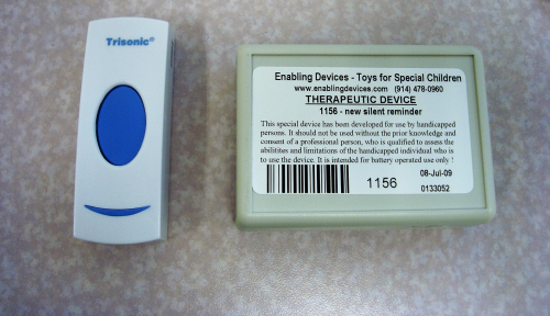 Two assistive communication switches on a tabletop: a white handheld switch with a large blue button and a rectangular therapeutic device with a printed label and barcode.