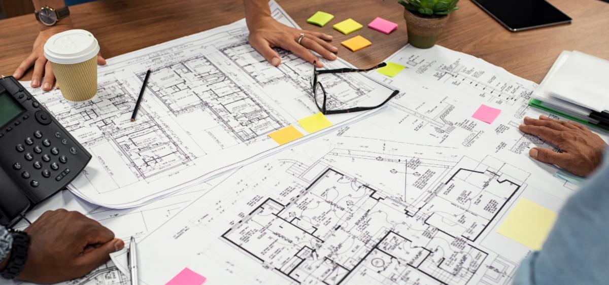 Hands of different people stretched around edges of blueprints on a table.
