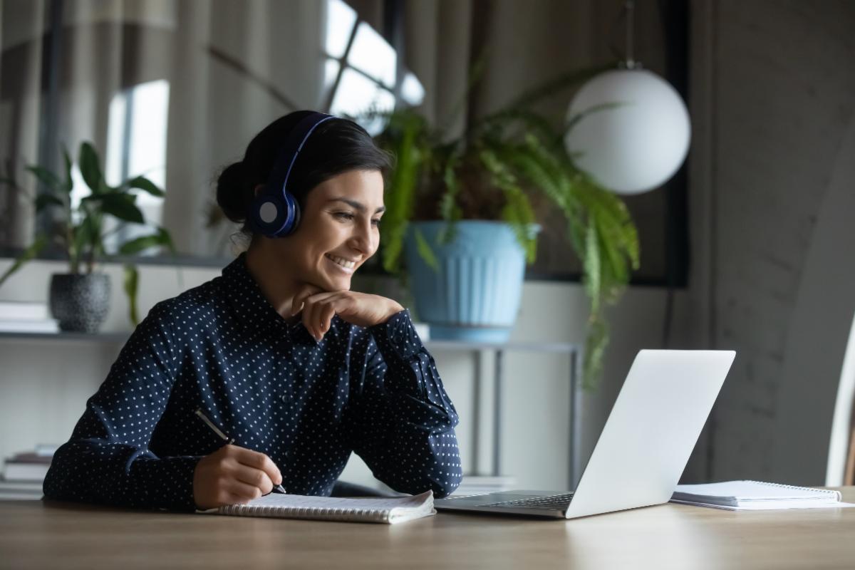 Woman with headphone listening to meeting on her laptop and taking notes.