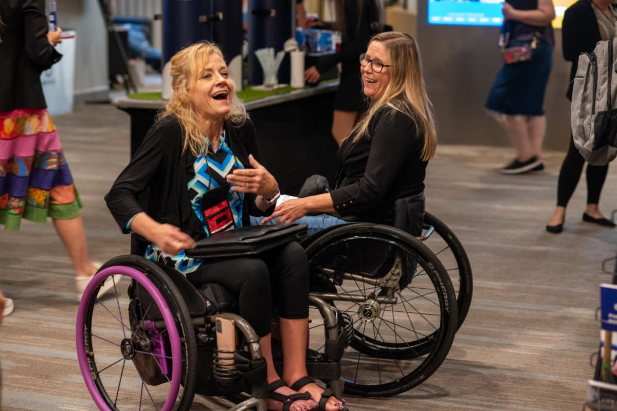 Two women who use wheelchairs laughing at a break in the National ADA Symposium.