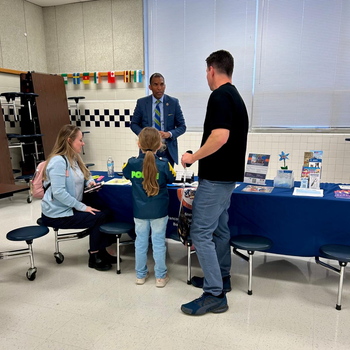 Family speaks with staff member at a brochure-filled information table in a school cafeteria, child wearing a “POLICE” jacket.