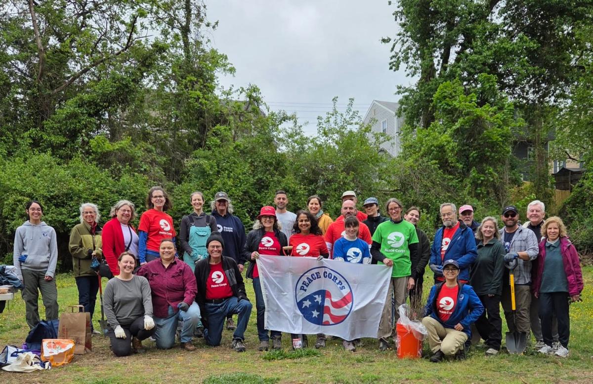 Group of adults pose in a field with trees while holding "Peace Corps" flag.