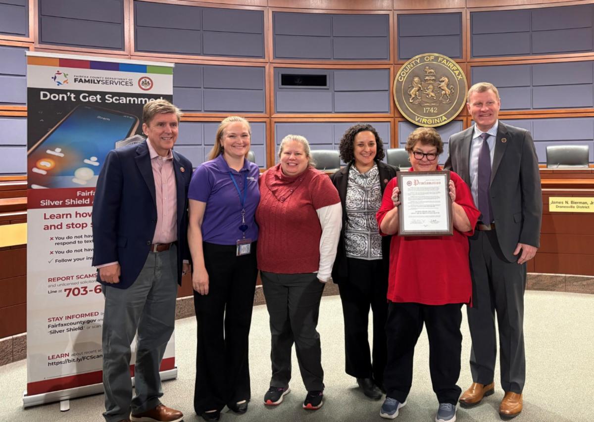 Group poses in Fairfax County auditorium as a woman holds a framed proclamation beside a “Don’t Get Scammed” banner.