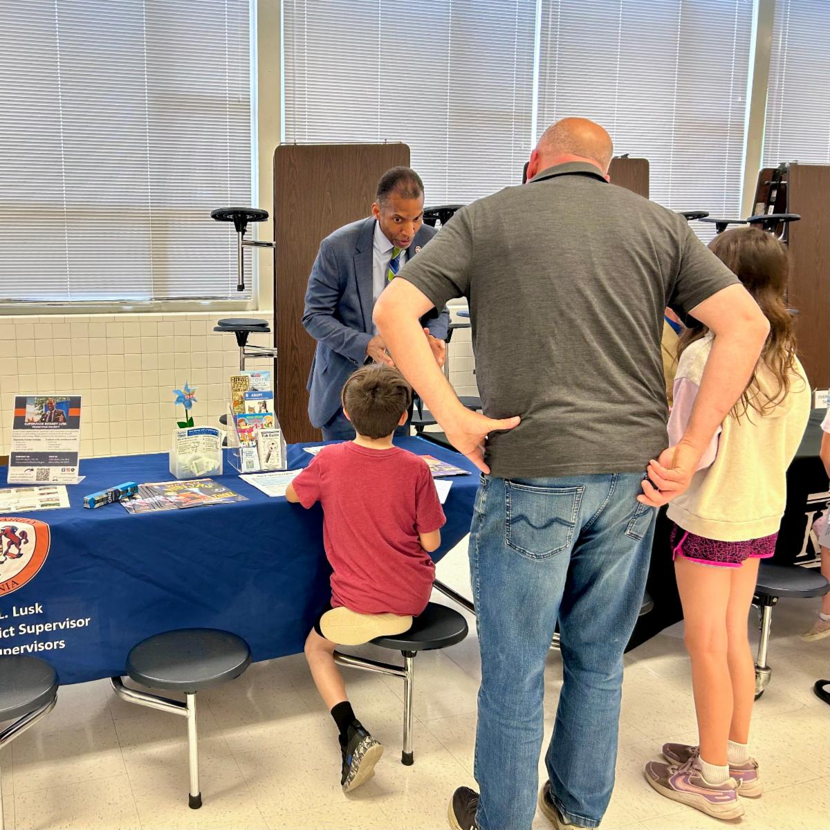Man and two children talk with a staff member at an information table covered in brochures and flyers in a school cafeteria.