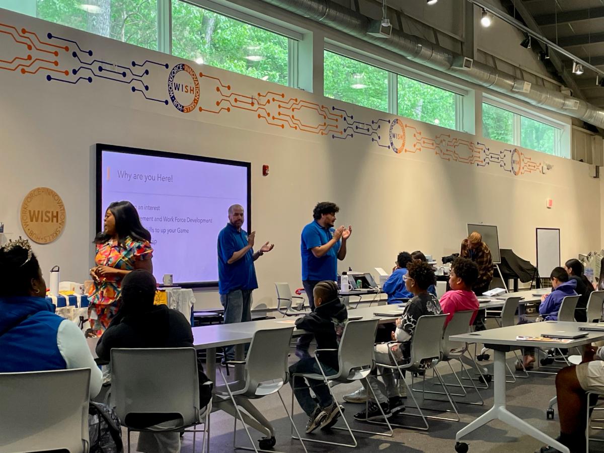 Facilitators lead a workshop at WISH hub while students seated at tables listen to a presentation on a large screen.