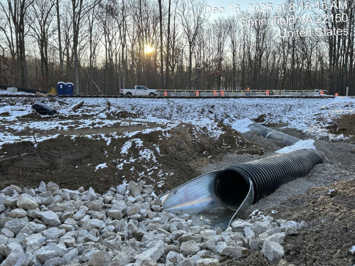 Snowy construction site with exposed drainage pipes, rocks, and a nearby road lined with cones and a work truck.
