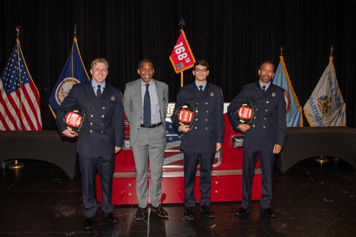 Three adults in firefighter uniforms pose with one suited adult in front of flags, one reading "168th."
