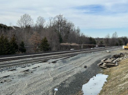Railroad tracks beside a gravel construction area with stacked timbers and a puddle, bordered by winter trees.