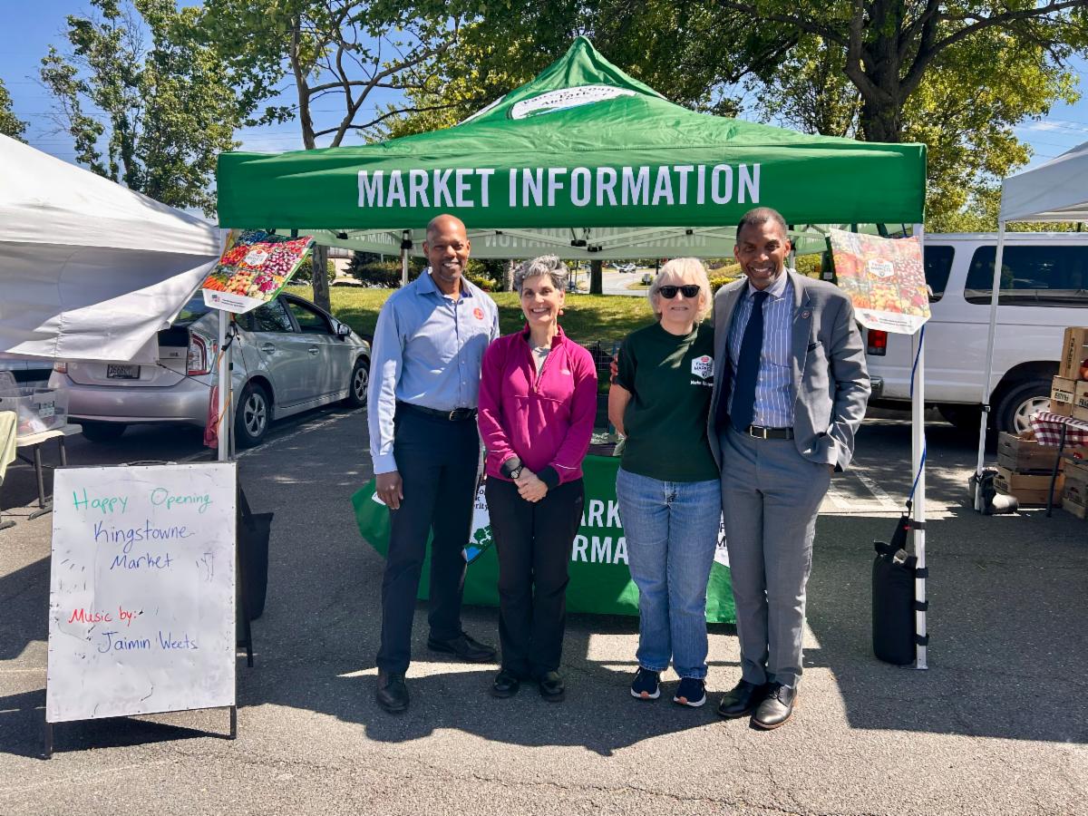 Four people stand in front of a green “Market Information” tent at the Kingstowne farmers market opening.