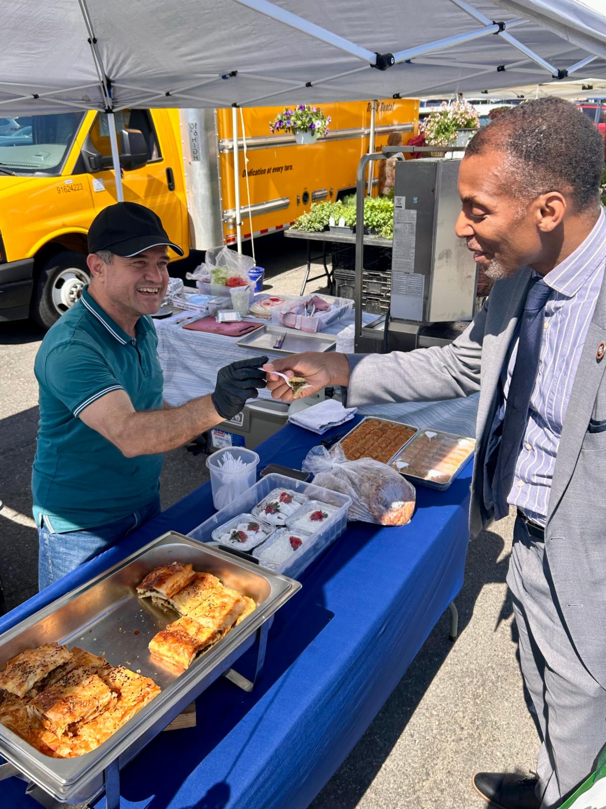 Vendor hands a food sample to a suited customer at a farmers market pastry stand under a white canopy.