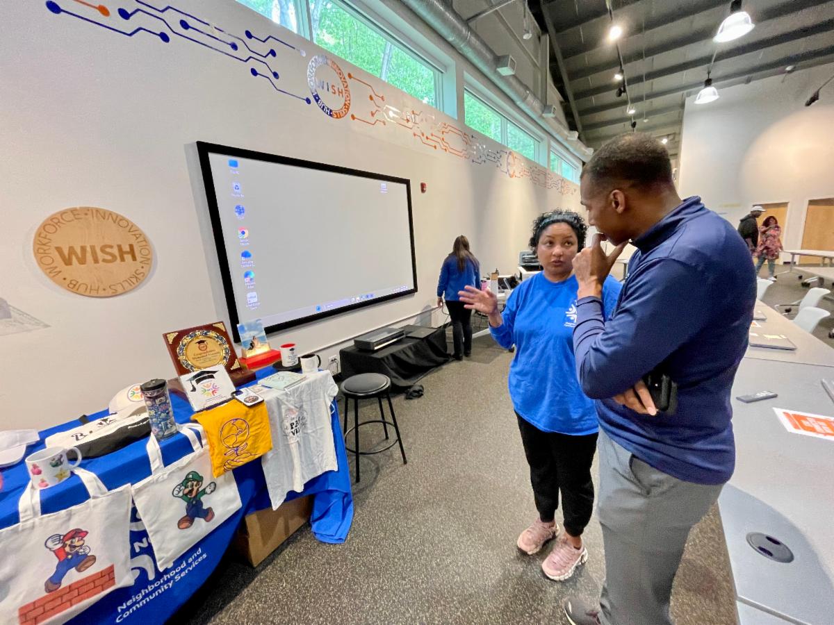 Two people talk near a WISH presentation table displaying custom tote bags, shirts, mugs, and awards in a classroom.