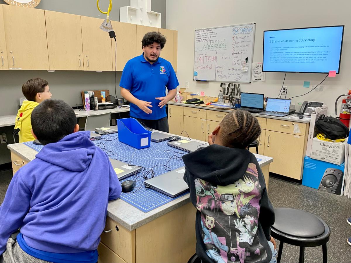 Instructor explains 3D printing stages to children seated around a lab table with laptops and a wall screen.
