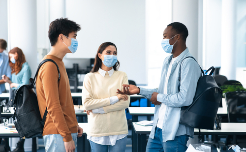 Healthcare_ Education_ Lifestyle And People Concept. Group of smiling diverse international students wearing protective medical masks and talking_ standing in lecture hall at the university