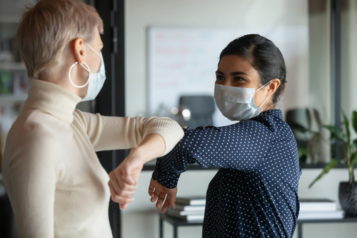 Smiling diverse female colleagues wearing protective face masks greeting...