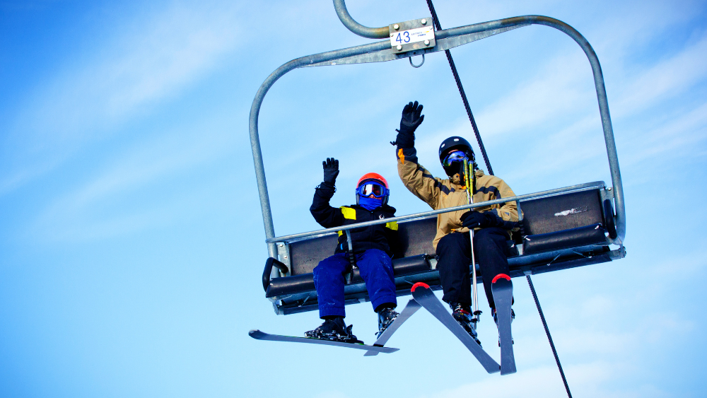 Two people in ski gear waving from a chair lift