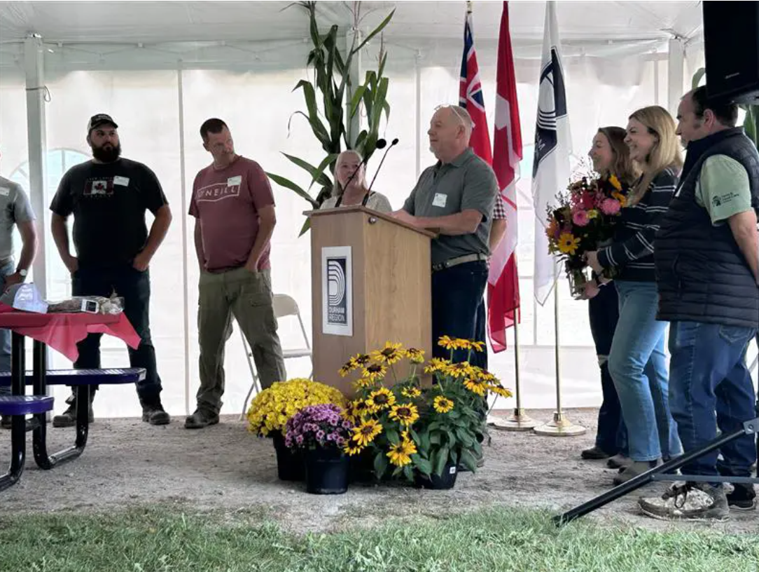Man speaks at a podium with people standing on both sides beneath a white tent.