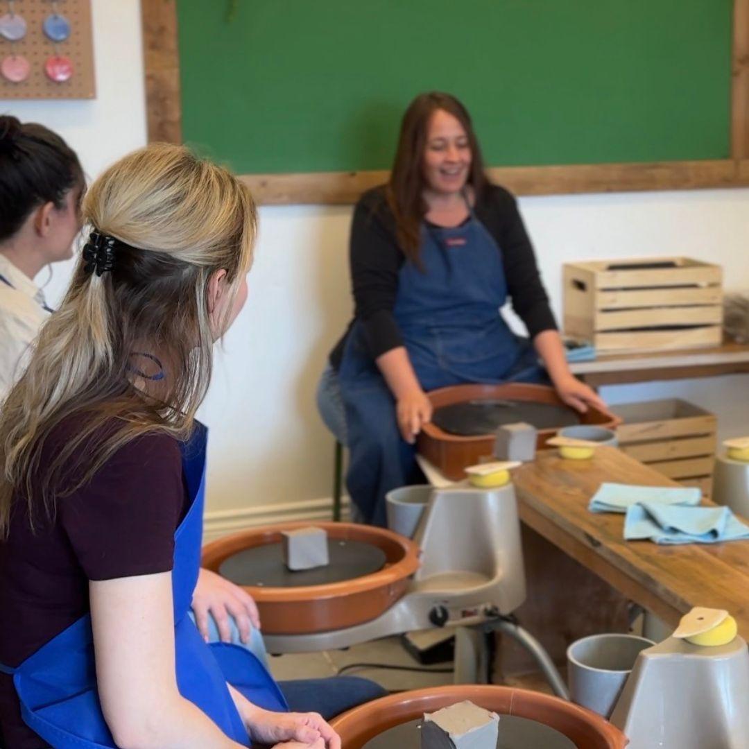 woman sits at potting wheel with students seated in front of her