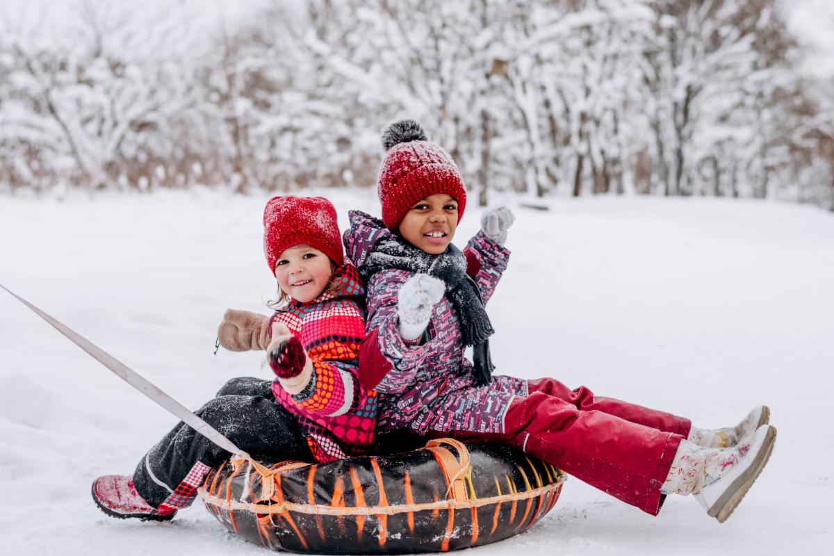 Two young children dressed in colourful snow suits are sitting on a tube sled in a snowy field