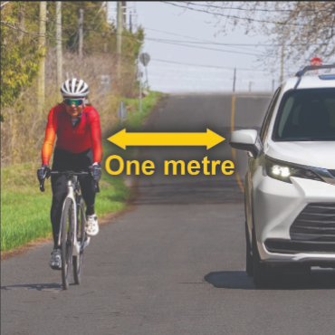Road safety image of cyclist riding left side of rural road as white car passes on right with one metre distance shown by double arrow trees poles and narrow verge in background reminding drivers to give cyclists space