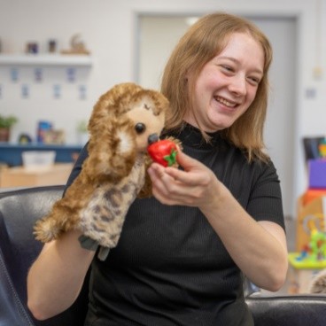 Registered Early Childhood Educator holding a puppet that is eating a plastic strawberry that they are holding in their other hand.