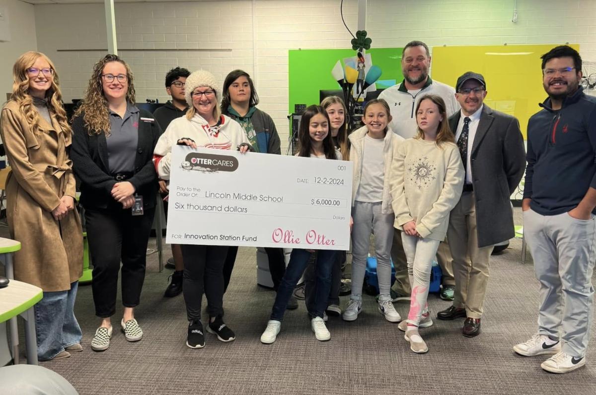 A dozen students and adults pose for a photo in a middle school classroom while holding a large novelty check from OtterCares for $6,000 for a 3D printing project. The check is signed by "Ollie Otter," dated 12-2-24, and made out to Lincoln Middle School.