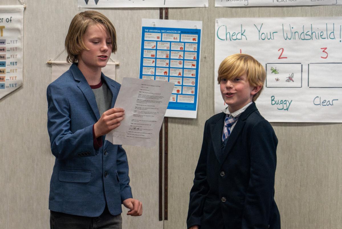 Dos niños de escuela primaria vestidos con chaquetas de traje hablan ante una multitud en su escuela.