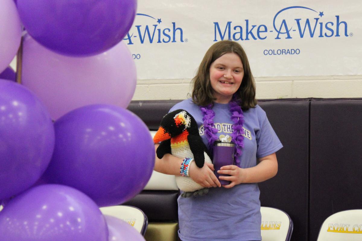 A middle school girl wearing a light purple T-shirt and holding a stuffed penguin smiles at an unseen crowd while standing on stage at a high school assembly. A tower of purple balloons are pictured to the left.