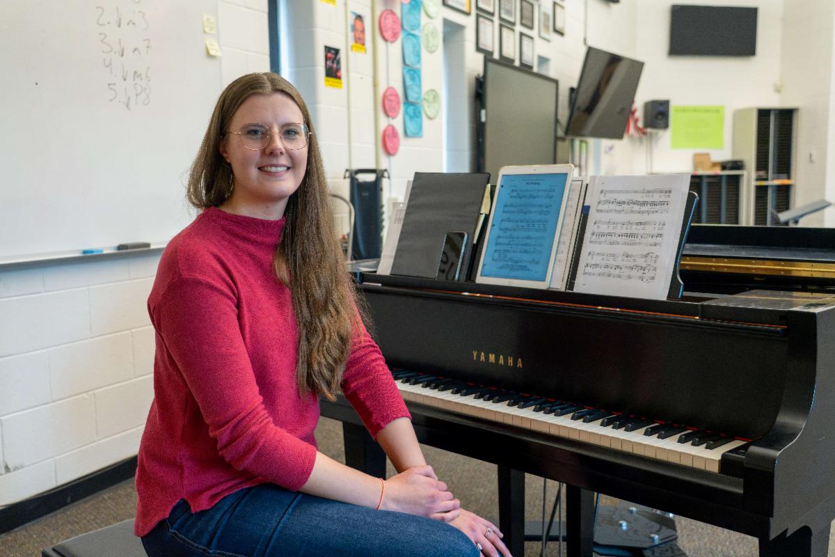 Portrait photo of a woman seated at a piano in a classroom.