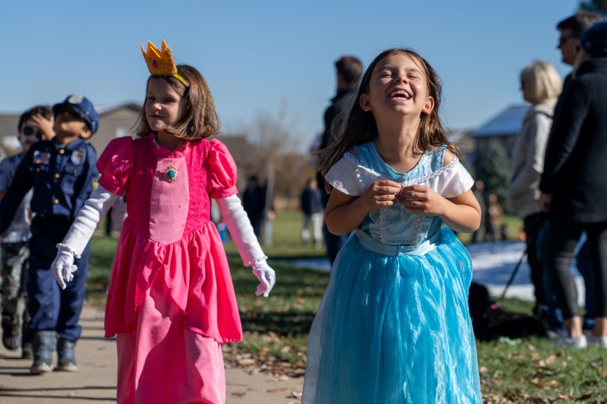 Two elementary school girls laugh and smile while dressed as princesses marching in a Halloween parade.
