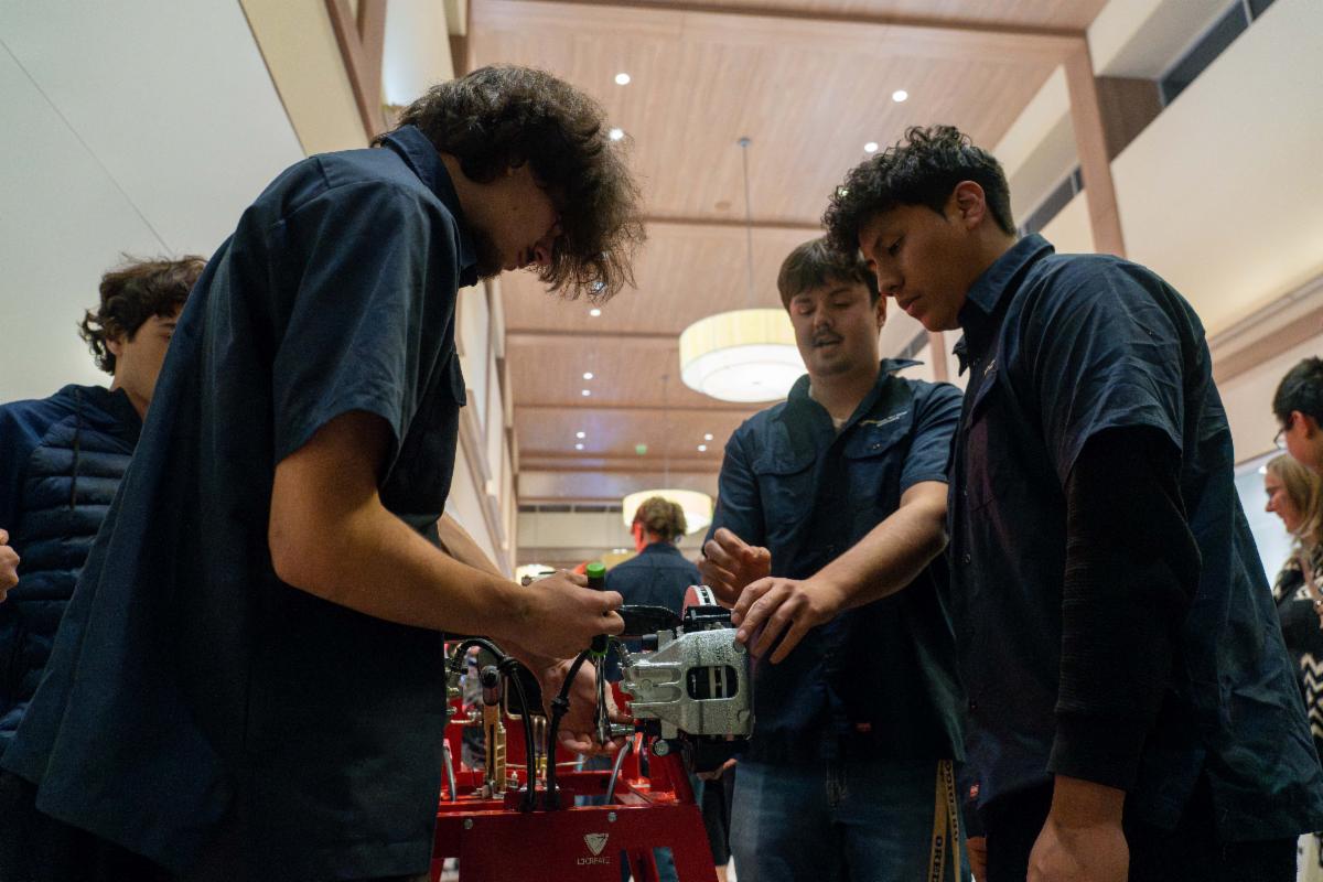 Cuatro jóvenes se encuentran en el pasillo de un centro comercial y rodean el motor de un automóvil. Al fondo se encuentran otros departamentos de educación profesional técnica, todos en stands mostrando sus proyectos finales.